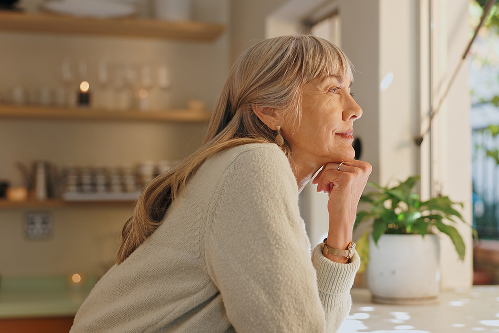 Mulher idosa sorrindo e olhando pela janela com expressão reflexiva, representando o cuidado com a saúde e a valorização da vida no fim do ano.